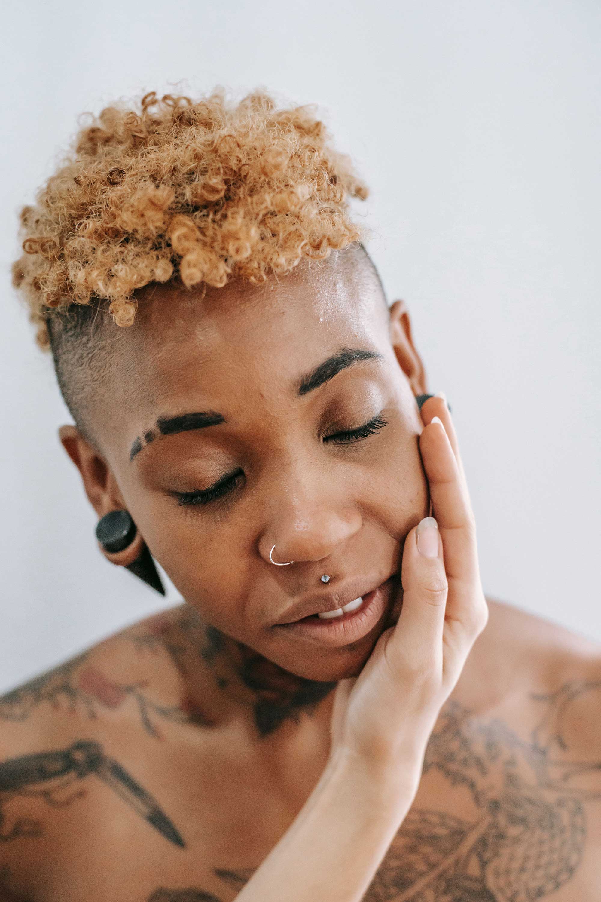 A person with short curly hair, facial piercings, and tattoos with eyes closed and hand gently resting on their face in a serene, introspective moment