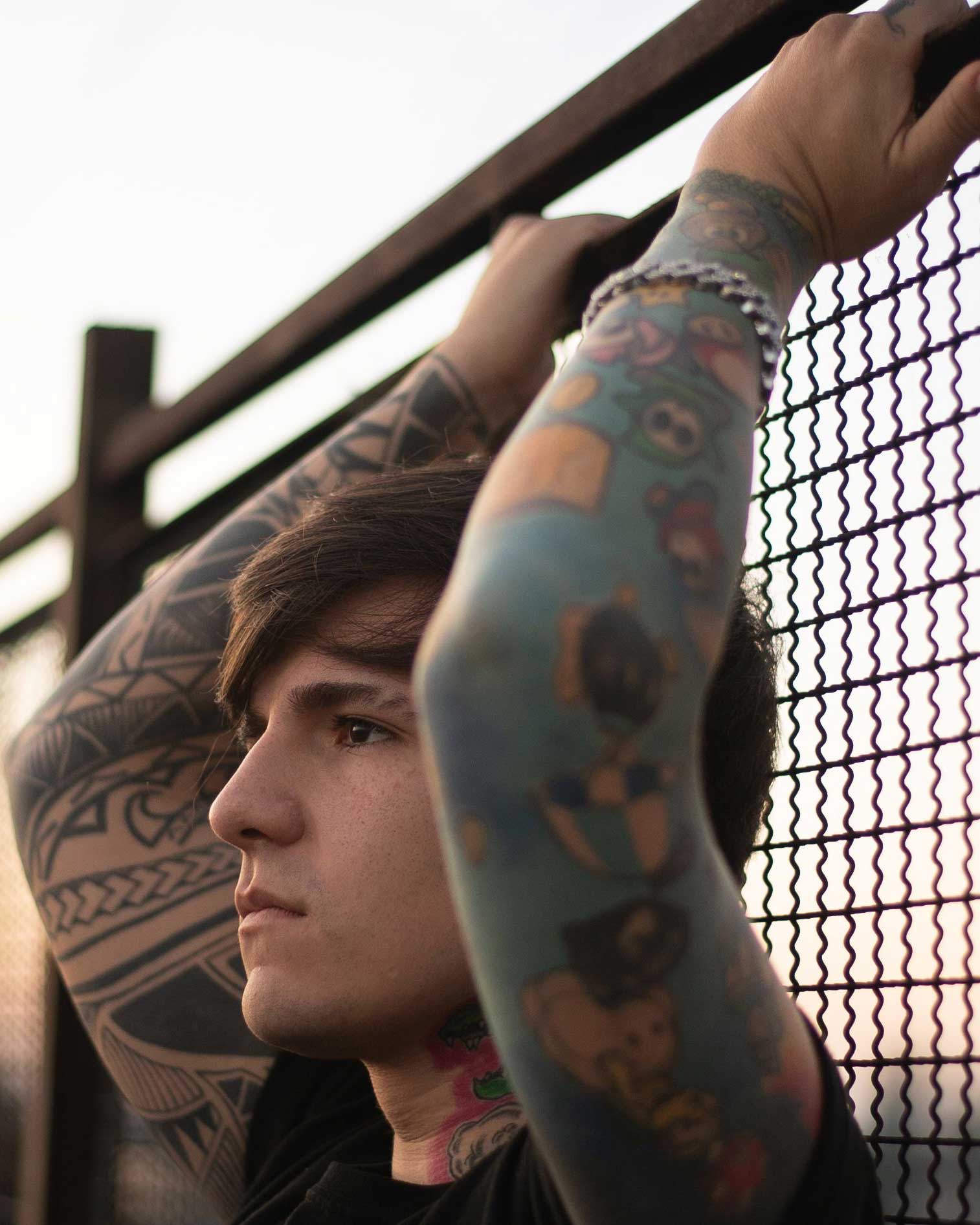 A person with long dark hair and full-sleeve tattoos standing with arms raised, gripping a fence and looking to the side with a contemplative expression