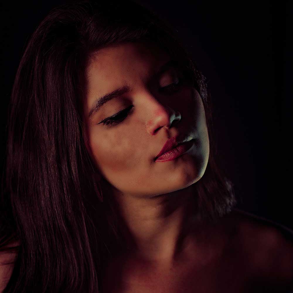A close-up portrait of a woman with long dark hair and red lips, eyes closed, in dramatic moody lighting