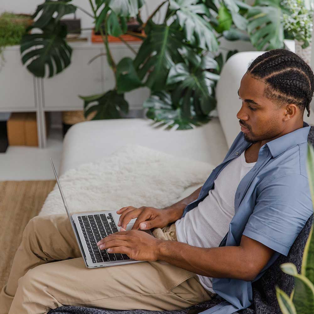 A person with braided hair sitting comfortably at home on a laptop, surrounded by lush houseplants