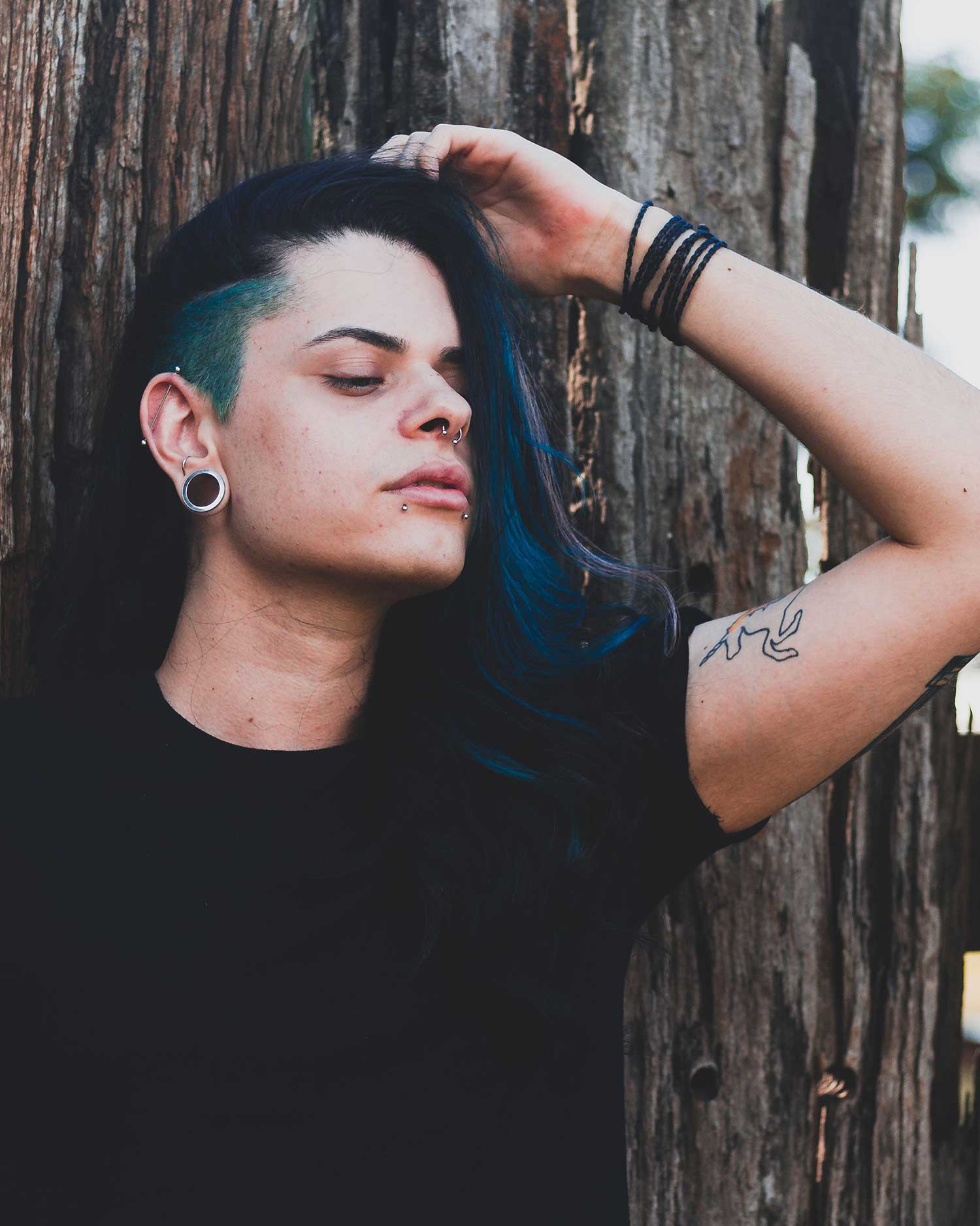 A person with long blue-dyed hair, ear gauges, and multiple piercings leaning against a weathered wooden wall with eyes closed, tattoos visible on their arm