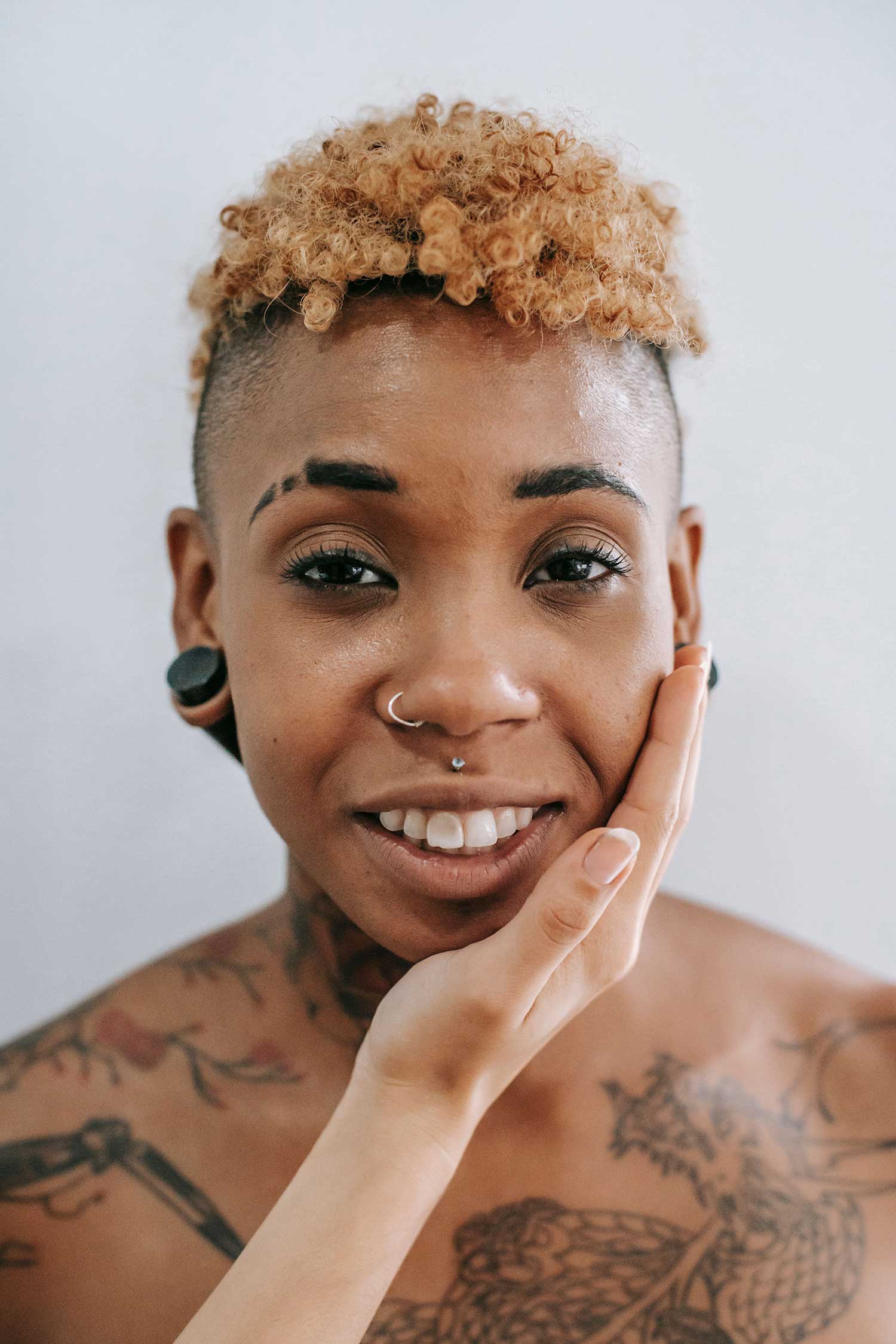 A person with short curly hair, facial piercings, and tattoos smiling softly while touching their face