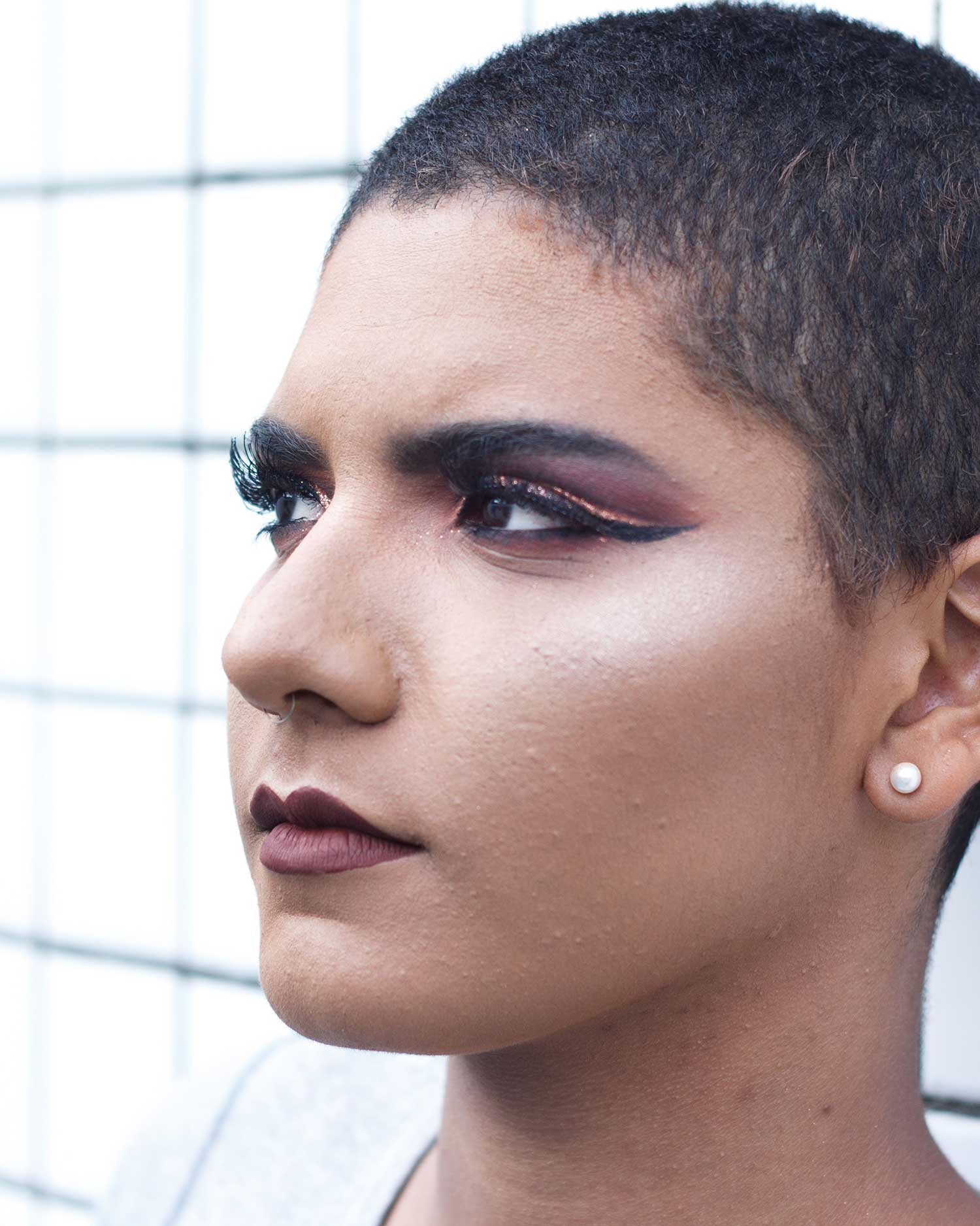A side profile portrait of a person with a shaved head and bold makeup including dark lipstick and dramatic eye makeup, looking thoughtfully to the side
