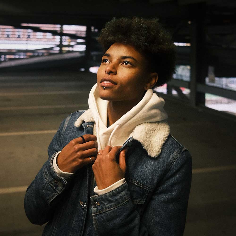 A person with natural curly hair and a nose piercing wearing a white hoodie and denim jacket, looking upward with a thoughtful expression in an urban setting