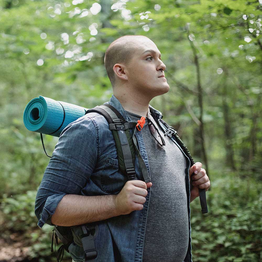 A person with a shaved head wearing a denim shirt and carrying a hiking backpack with a rolled mat, standing in a lush green forest and looking upward