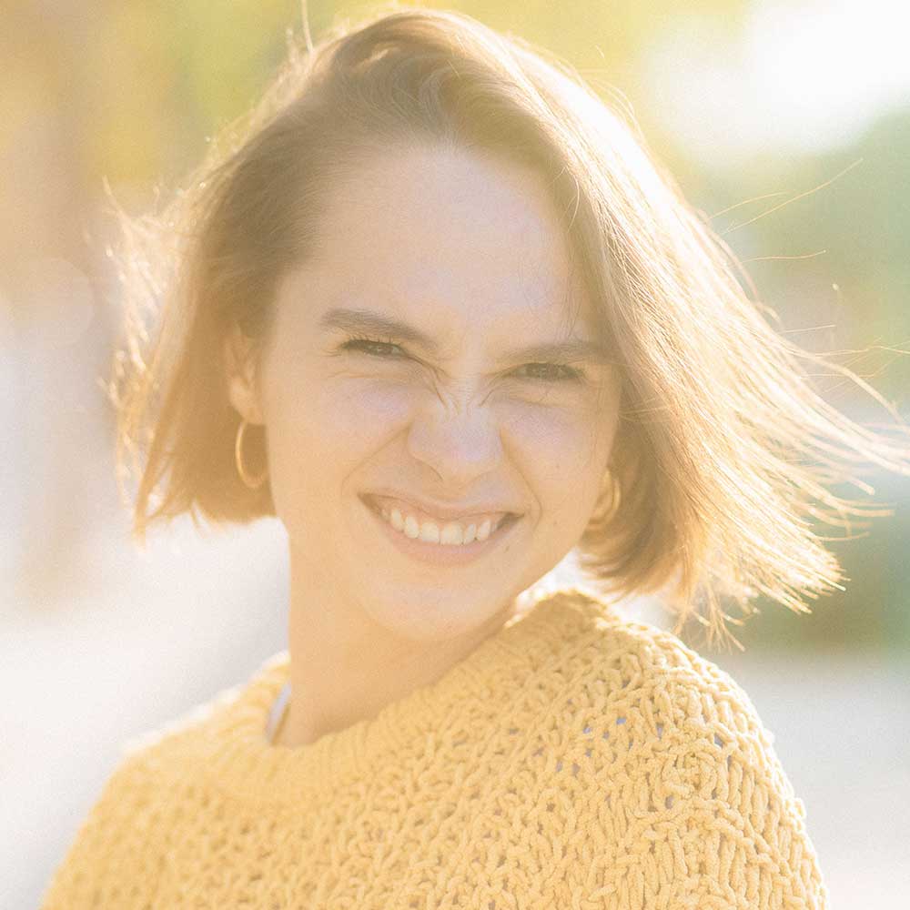 A woman with short reddish-blonde hair smiling warmly in bright sunlight, wearing a yellow knit sweater