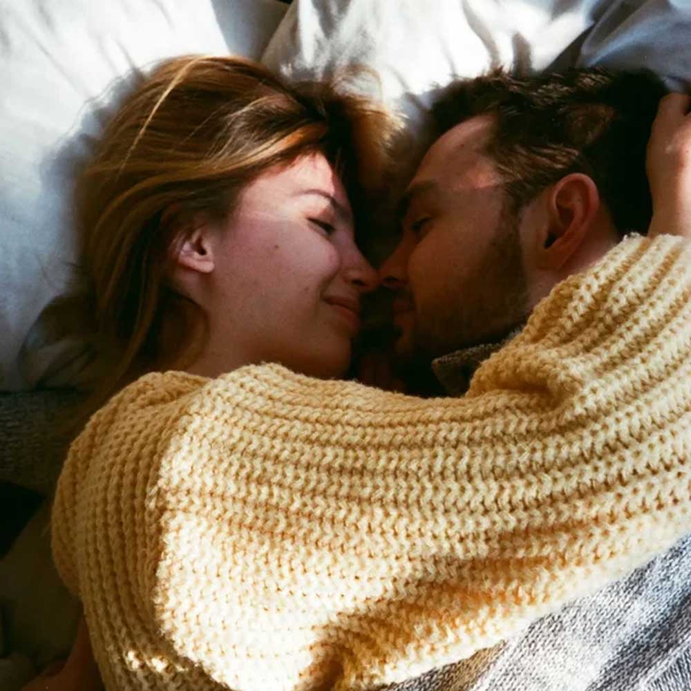 Two people lying close together in bed, faces nearly touching and smiling softly at each other in warm light