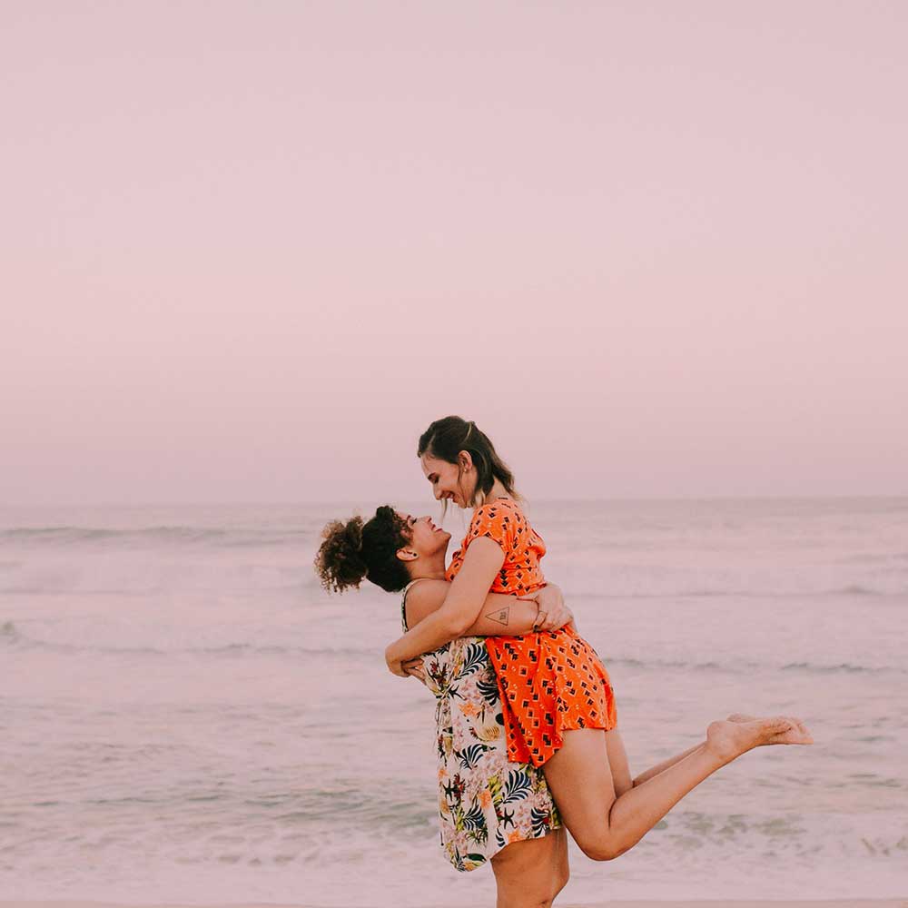 Two people laughing joyfully on a beach at sunset, one lifting the other in a playful, loving embrace